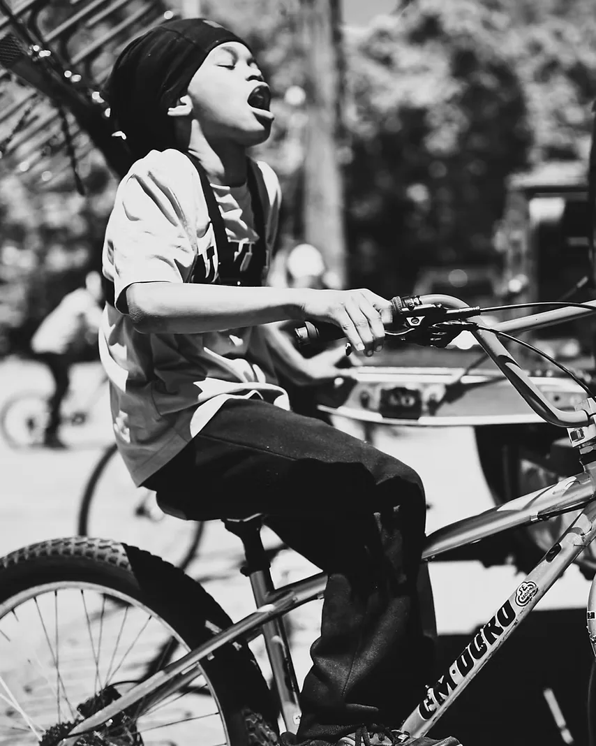 A young rider on a BMX bike at a ride-out, head thrown back mid-shout.