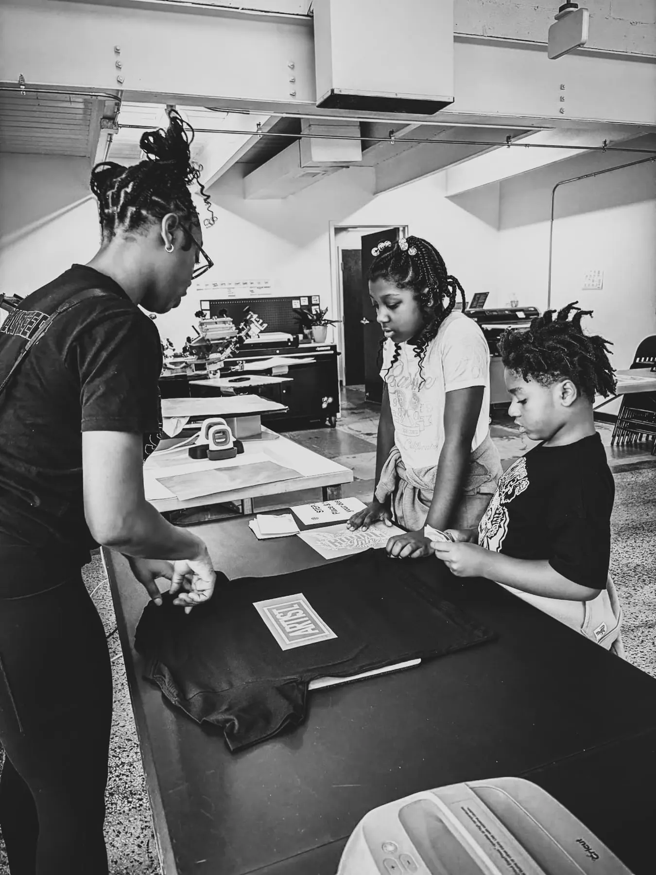 Young people gathered around a table at a Mount Pleasant Printing session, looking over a freshly printed shirt.