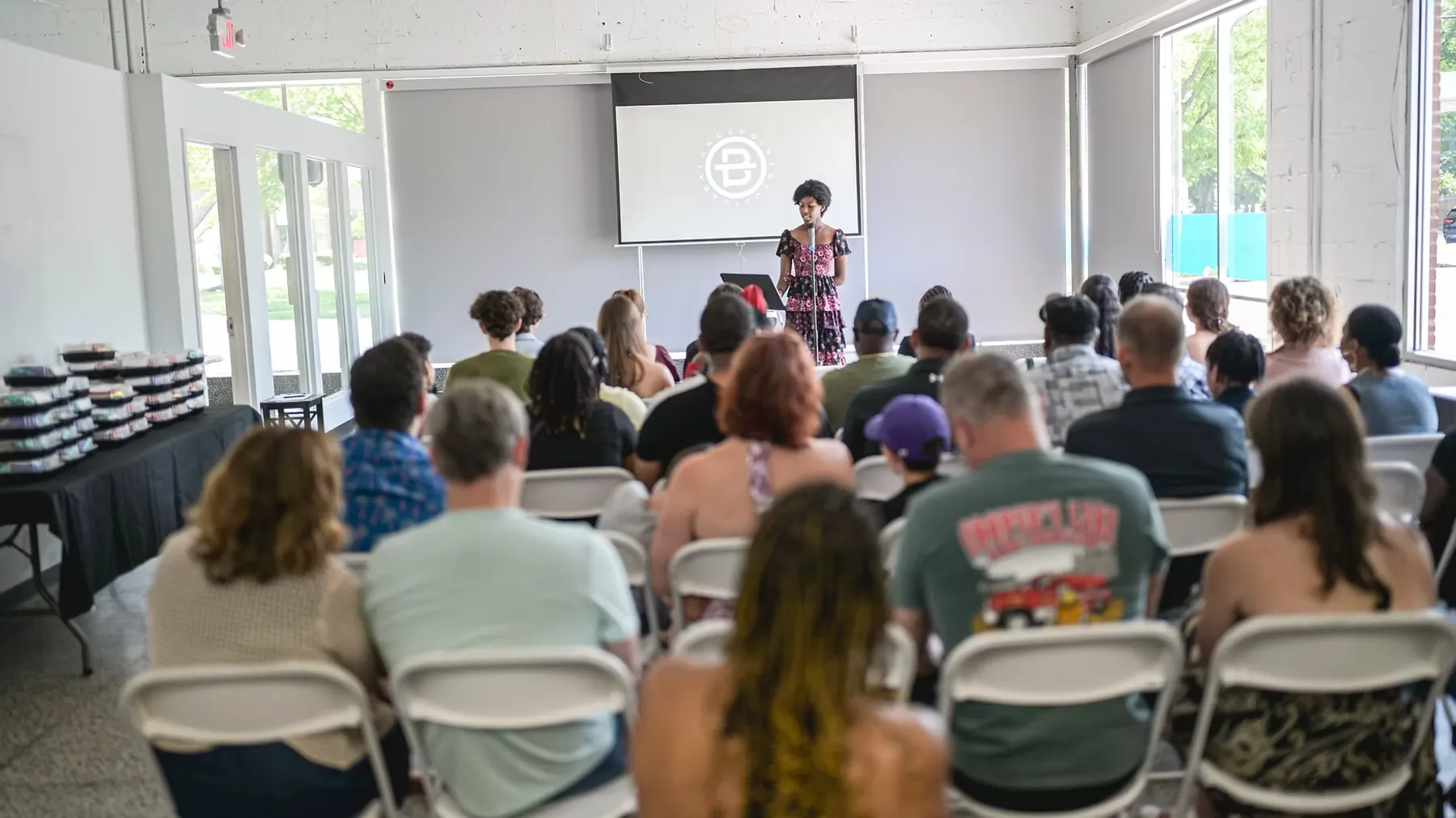 A young woman reads at a microphone to a full room of seated guests.