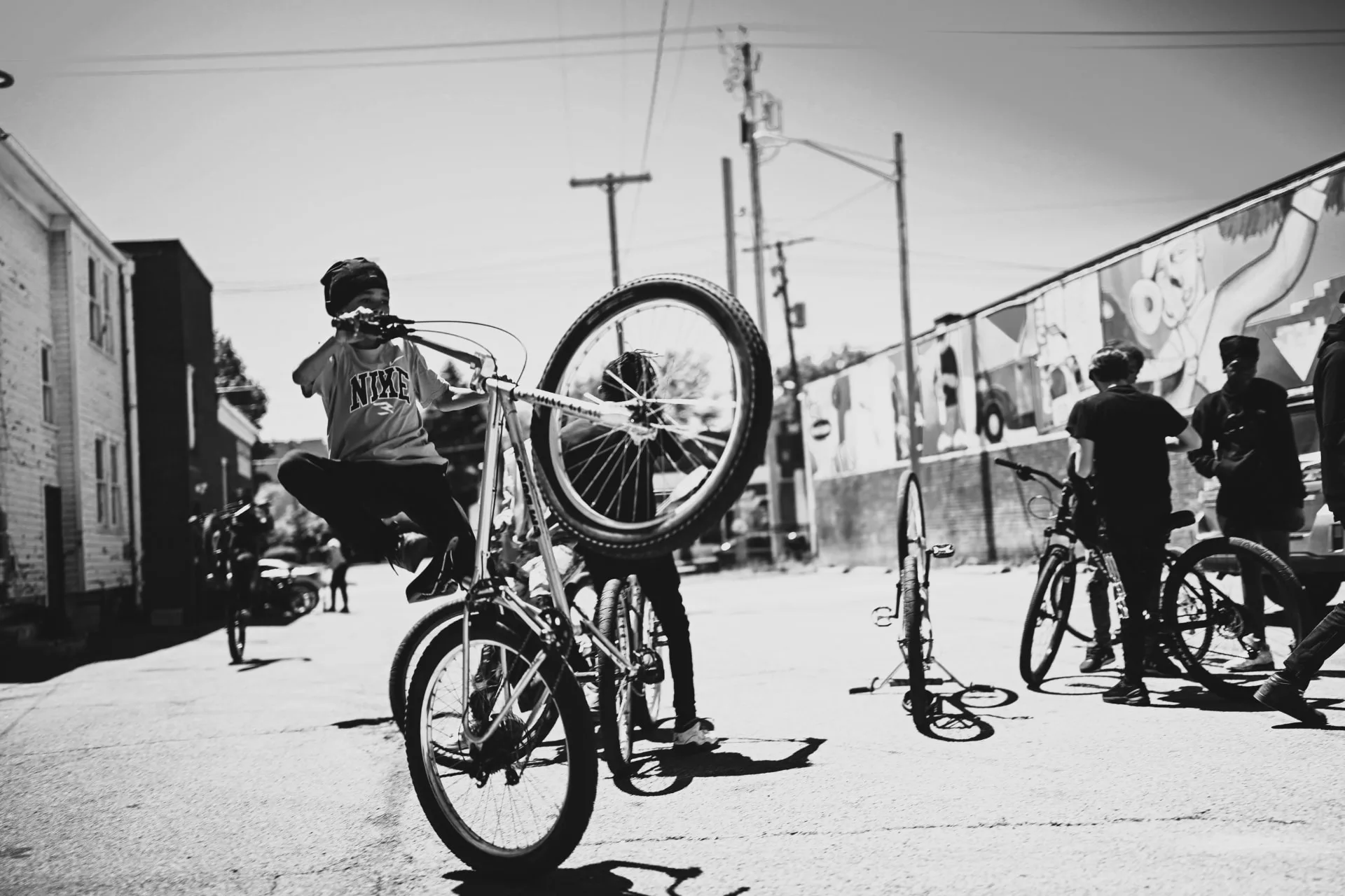 A young rider pops a wheelie on a BMX at a ride-out, front wheel high, friends and murals behind.